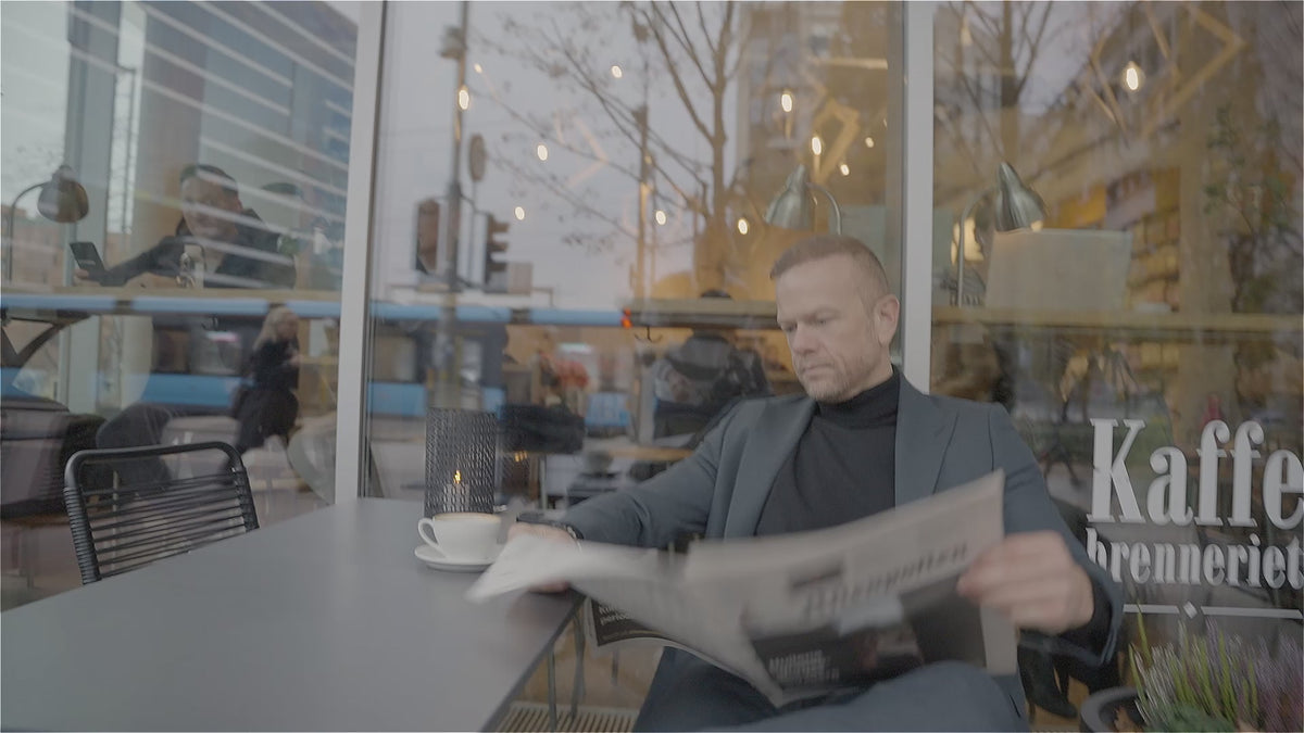 Mann i grå dress leser avisen ved et kafébord i byen