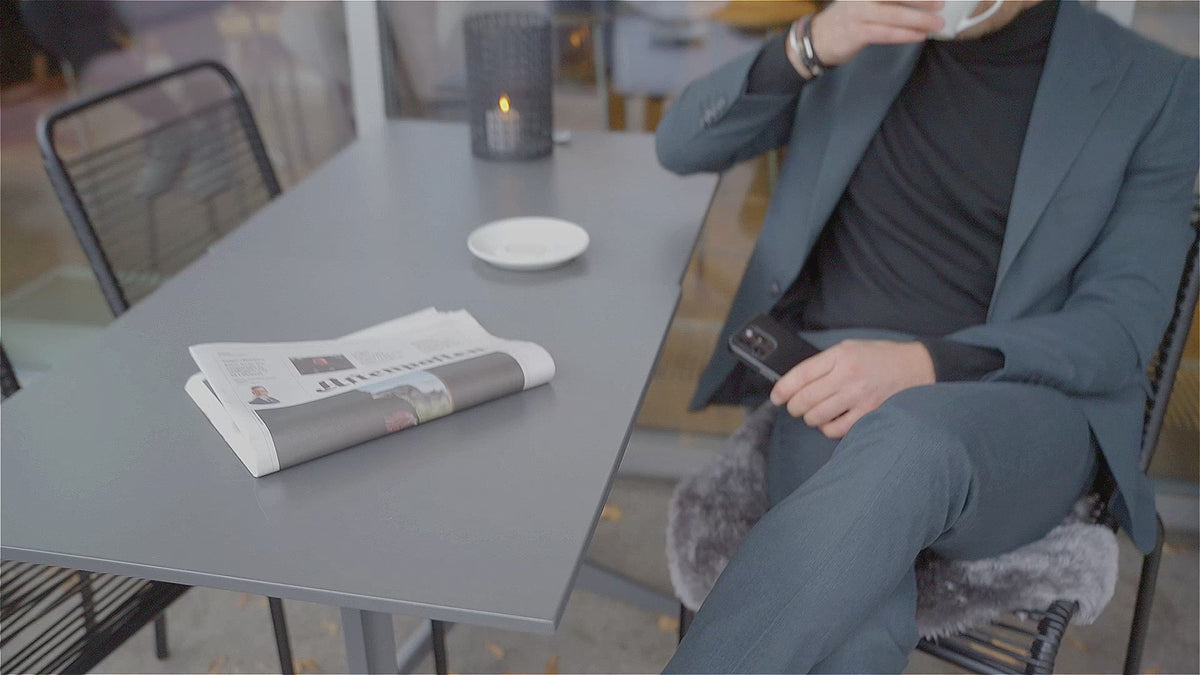 Mann i grå dress og sort finstrikk sitter ved et kafébord med kaffekopp og avis